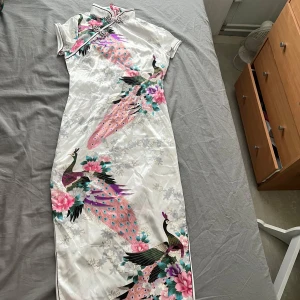 Klänning med påfåglar slits 2 sidor - Elegant vit långklänning i siden med färgglada påfåglar och blommor. Klänningen har korta ärmar och en hög krage med knäppning. Perfekt för en speciell tillställning.