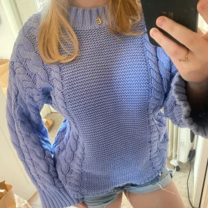 Ljuslila stickad tröja - Säljer en mysig ljuslila stickad tröja med flätstickade detaljer längs ärmarna och sidorna. Tröjan har en rund halsringning och långa ärmar, perfekt för kyligare dagar. Passar bra till jeans eller kjol för en avslappnad look.