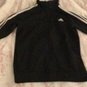 Svart träningsjacka från Adidas - Säljer en svart träningsjacka från Adidas med vita ränder längs ärmarna och dragkedja framtill. Jackan har en hög krage och det klassiska Adidas-loggan på bröstet. Perfekt för träning eller en sportig look.