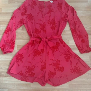 Rosa blommig jumpsuit med bälte - Supersöt rosa jumpsuit med blommigt mönster, långa ärmar och knytbälte i midjan. Klänningen har dragkedja i sidan och är perfekt för dig som gillar färg och detaljer. Flowy material och snygg passform. Använde bara en gång. Förminskad i storleken som hm storlekar  brukar vara. Är mer som M storlek. Bröst 102cm, midjan 85cm, längd 82cm.