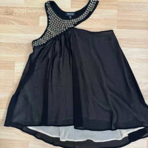 Svart  tunika med paljettdetaljer Topshop - Supersnygg svart tunika från Topshop med glittriga paljettdetaljer vid halsringningen. Tunika är ärmlös och har ett flowy, lätt tyg i polyester med ljus innerklänning. Perfekt för dig som vill sticka ut med en cool och elegant look. Använd en gång.