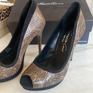 Bruna klackar  med ormskinnsmönster Kenneth Cole - Snygga klackar från Kenneth Cole i brunt ormskinnsmönster med svarta detaljer. Skorna har hög klack, öppen tå och plattform. Utsidan är i skinn med struktur och insidan är svart. Aldrig använt tidigare. Kartong finns till, 12 cm