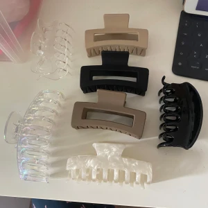 Hårklämmor - Säljer ett set med sju stora hårklämmor i olika färger och material. Färgerna är svart, beige, genomskinlig, pärlvit och iriserande. Klämmorna har rektangulär och klassisk form, perfekta för att snabbt sätta upp håret. Stilrena och trendiga accessoarer till håret.❤️