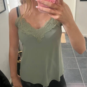 Olivgrön linnetopp med spetsdetaljer - Supersnygg olivgrön linnetopp med tunna axelband och v-ringning. Toppen har fina spetsdetaljer längs ringningen både fram och bak. Perfekt att styla med jeans eller kjol för en trendig look.