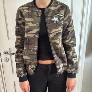Camouflage bomberjacka med patches - Säljer en snygg bomberjacka i camouflage med coola patches på bröstet – stjärna, vingar och krona. Jackan har dragkedja, fickor och muddar vid ärmslut och midja. Perfekt för dig som vill sticka ut med streetstyle-vibe.
