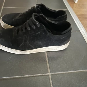 Svarta arigatos i mocka - Svarta sneakers från arigato med ovandel i mocka och klassisk vit sula. Skorna har snörning och en stilren, låg profil som passar till det mesta. Perfekta för dig som gillar en enkel och clean look.