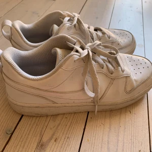 Vita Nike sneakers i skinn, stl 40 - Fräscha vita Nike sneakers i skinn med klassisk design och perforerad tå. Skorna har snörning och en platt sula, perfekt för en clean och sportig look. Bekväm mesh på insidan och diskret Nike-logga på plösen.