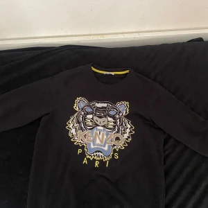 Svart Kenzo tröja med tigertryck - Svart långärmad tröja från Kenzo med ikoniskt broderat tigerhuvud i blått, gult och grått framtill. Rund halsringning med gul detalj på insidan. Perfekt för dig som vill sticka ut med en streetstyle-look.