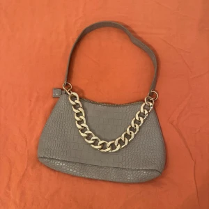 Grå/beige axelväska med kedjedetalj - Snygg grå/beige axelväska i croco-mönstrat syntetmaterial med en chunky guldfärgad kedja framtill. Väskan har dragkedja upptill och ett innerfack med dragkedja. Perfekt storlek för det viktigaste och ger en cool vibe till din outfit.