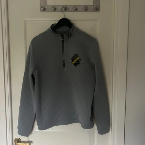 Grå quiltad tröja AIK x FootJoy - Quiltad grå långärmad tröja från FootJoy med AIK-logga på bröstet och FJ-detalj på kragen. Tröjan har half zip och hög krage, perfekt för kyliga dagar. Snygg och sportig stil som sticker ut på golfbanan eller i stan.