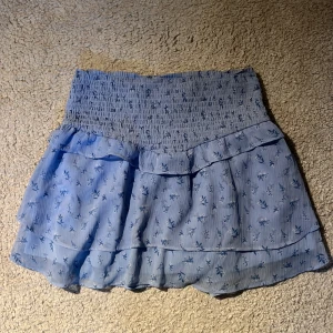 Blommig blå volangkjol Gina Tricot - Supersöt ljusblå kortkjol från Gina Tricot med blommigt mönster och volanger. Kjolen har smock i midjan för en bekväm passform och är gjord i ett lätt, flowy material som passar perfekt till varma dagar💗Knappt använd!! 