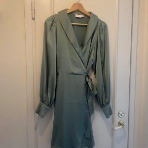 Ljusgrön omlottklänning från Vila - Supersnygg ljusgrön omlottklänning från Vila i kort modell. Klänningen har långa puffärmar med breda manschetter och knappar, markerad krage och knytband i midjan. Materialet är mjukt och har en lätt glansig finish som ger en elegant känsla.