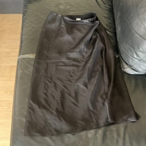 Svart långkjol i satin - En stilren svart långkjol i glansigt satinmaterial. Kjolen har en rak och enkel design som ger en clean look. Perfekt för dig som gillar minimalistisk stil och vill ha något som känns både trendigt och klassiskt.