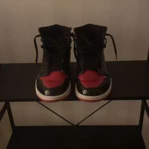 Nike Air Jordan 1 Bred Highs - Säljer ett par ikoniska Nike Air Jordan 1 Highs i svart och rött med vita sulor. Skorna har klassisk snörning, rund tå och är tillverkade i skinn med glansig finish. Perfekta för dig som vill sticka ut med en tidlös sneaker med streetkänsla.