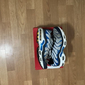 Nike Air Max Plus blå/vit sneakers - Nike Air Max Plus sneakers i blått, vitt och svart med ikoniska vågmönster och Tn Air-detalj på hälen. Ovandel i mesh och syntet, synlig Air Max-dämpning och snörning. Perfekta för dig som gillar streetwear och sportig stil.