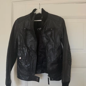 Svart skinnjacka från Conbipel - Svart skinnjacka från Conbipel med coola quiltade detaljer på armbågarna, ribbad krage och muddar. Jackan har flera fickor med dragkedja och tryckknapp, samt snygga sömmar som ger en schysst form. Perfekt för dig som vill ha en edgy look.