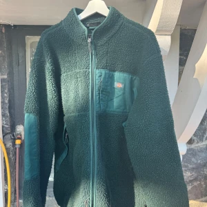Mörkgrön fleecejacka från Dickies XL - Mysig mörkgrön fleecejacka från Dickies i storlek XL. Jackan har hel dragkedja, förstärkta partier på bröst och armbågar samt en liten logga på bröstet. Perfekt för kalla dagar och har en riktigt skön teddylook.