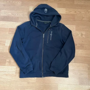 Mörkblå hoodie från Sail Racing - Mörkblå hoodie från Sail Racing med dragkedja och broderad logga på huvan. Tröjan har långa ärmar, huva och en clean design med Sail Racing-tryck på bröstet. Perfekt för dig som gillar sportig och avslappnad stil.