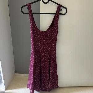 Vinröd blommig klänning  - Vinröd kortklänning från Abercrombie & Fitch med små blommor, markerad midja och utställd kjol. Klänningen har ärmlös design och snygg öppen rygg med twistad detalj. Perfekt för dig som gillar en feminin och trendig look.