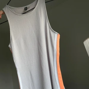 Grå sportig ribbad klänning - Grå ribbad ärmlös klänning med stretchig passform. Klänningen har orangea paneler längs sidorna som ger en sportig vibe. Materialet är stretchigt och bekvämt, perfekt för varma dagar. Enkel och stilren design med rund halsringning.