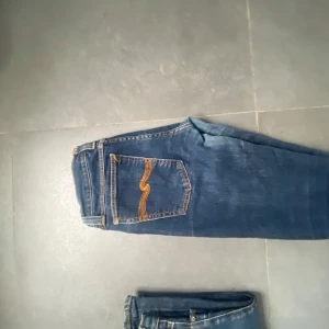 Mörkblå jeans från Nudie Jeans - Snygga mörkblå jeans från Nudie Jeans med klassiska orangea sömmar och detaljerad bakficka. Jeansen har fem fickor, knappgylf och är tillverkade i slitstarkt denim. Perfekta för dig som gillar en tidlös och clean look.