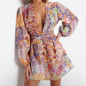 Färgglad omlottklänning med mönster - Superfin kort omlottklänning med djup v-ringning och långa puffärmar. Klänningen har ett färgstarkt patchworkmönster i lila, orange, rosa, grön och vit, samt knytband i midjan och öppen rygg. Perfekt för dig som vill sticka ut med en unik look!