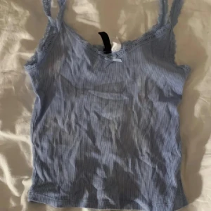 Ljusblå ribbad linnetopp med spets - Supersöt ljusblå linnetopp med tunna axelband och spetsdetaljer längs urringningen. Toppen är ribbad och har en liten rosett framtill. Perfekt för varma dagar eller som layering-piece.