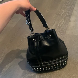 ZARA❤️Svart bucket handväska med nitar - Cool svart bucket handväska i skinnimitation med silvriga metalldetaljer och kedjehandtag. Väskan har snörning upptill och dragkedja med nitar längst ner för en edgy look. Perfekt storlek för allt du behöver och riktigt snygg att bära till vardagsoutfiten.