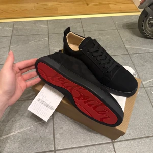 Svarta Louboutin sneakers med röd sula - Snygga svarta sneakers från Christian Louboutin i mocka med klassisk röd sula och diskret logga på hälen. Skorna har rund tå, svarta snören och en stilren design som sticker ut med den ikoniska röda undersidan.
