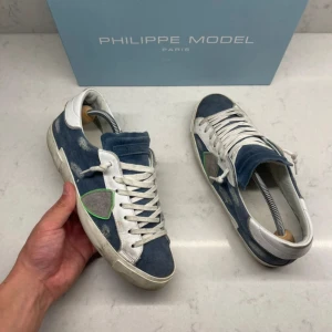 Philippe Model blå sneakers med mockadetaljer - Snygga sneakers från Philippe Model i blått tyg med vita skinnpaneler och detaljer i mocka. Skorna har snörning, rund tå och en cool grön detalj på sidan. Perfekt för dig som gillar en avslappnad men trendig stil.
