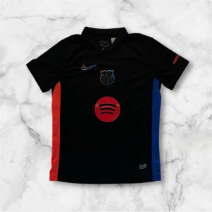 Svart FC Barcelona Nike fotbollströja - Snygg svart FC Barcelona fotbollströja från Nike med klubbmärke och Nike-logga i rött och blått. Spotify-logga i rött på bröstet och UNHCR-tryck på ryggen. Tröjan har korta ärmar och färgdetaljer i rött och blått på sidorna. Tillverkad i lätt och ventilerande material.
