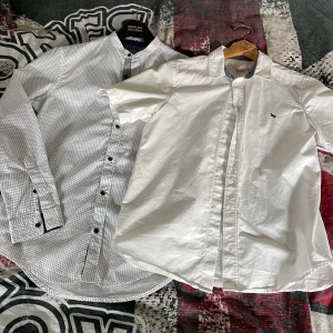 Två vita skjortor - Säljer två snygga skjortor: en vit kortärmad skjorta från Gant med liten logga på bröstet och en vit långärmad skjorta från KappAhl med diskret svart mönster och svarta knappar. Perfekta för en clean och fräsch look.