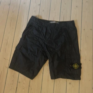Navy cargo shorts från Stone Island - Navy cargo shorts från Stone Island med klassisk patch på benet. De har flera fickor, inklusive sidofickor med knapp och cargofickor. Materialet är lätt, glansigt och prassligt. Snygg och avslappnad passform med midwaist. 