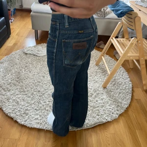 Wrangler raka jeans i mörkblå denim - Säljer ett par klassiska mörkblå jeans från Wrangler med raka ben och normal passform. Jeansen har fem fickor, snygga kontrastsömmar och läderpatch med logga bak. Perfekta för dig som gillar en tidlös look och vill ha ett par jeans som funkar till allt.