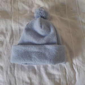 Ljusblå mössa med tofs från Monki - Supermjuk ljusblå mössa från Monki med bred uppvikt kant och en fluffig tofs på toppen. Perfekt för kalla dagar och enkel att matcha med olika outfits. Tillverkad i ett mjukt syntetmaterial som känns skönt mot huden. Oanvänt/nyskick, men sååå fin och skön. Pris går att diskuteras