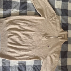 Beige halvzip tröja i lammull från GANT - Snygg beige tröja från GANT i superfin lammull. Halvzip-dragkedja och broderad logga på bröstet. Ribbstickade muddar och hög krage ger en clean och klassisk look. Perfekt för lager-på-lager under våren och sommar kvällar.