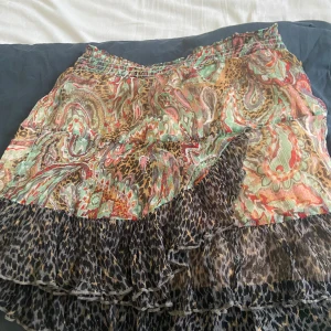 Mönstrad kortkjol från Alix - Färgstark kortkjol från Alix med mix av paisley och leopardmönster i grönt, rött, beige och brunt. Kjolen har volangkant nertill och resår i midjan för skön passform. Perfekt för dig som gillar unika prints och vill sticka ut.