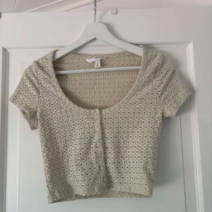 Beige croppad topp från H&M - Superfin croppad topp från H&M i beige med korta ärmar och rund halsringning. Toppen har ett hålmönster i tyget och knappar framtill. Perfekt för en chill och trendig look, lätt att matcha med jeans eller kjol.