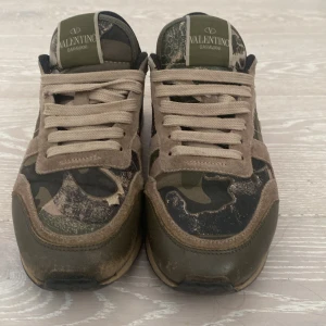 Valentino Garavani Rockrunner sneakers - Snygga Valentino Garavani Rockrunner sneakers med camouflage-mönster i grönt, beige och svart. Skorna har detaljer i mocka, skinn och textil, samt en röd hälkappa och nitar på sulan. Klassisk snörning och platt sula. Perfekt för dig som gillar streetstyle med lyxig touch.