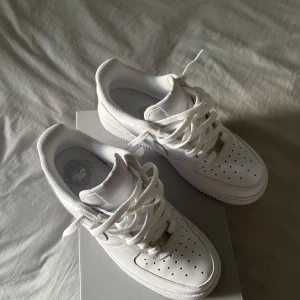 Nike Air Force 1 '07 helvita sneakers - Nike Air Force 1 '07 i helvitt skinn med klassisk låg siluett, perforerad tå och vit sula. Ikonisk Swoosh-logga på sidan och metallplatta vid snörningen. Perfekta för en clean och stilren look. Helt nya i kartong! 