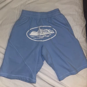 Blåa corteiz short - Blåa corteiz shorts med elastisk midja och ett stort vitt tryck framtill som föreställer ett fartyg. Shortsen har sidofickor och en bakficka, samt är tillverkade i ett mjukt och bekvämt material. Perfekta för sommardagar eller chill hemma.