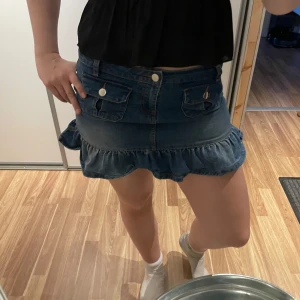Jeanskjol med volangkant - Säljer en blå jeanskjol med volangkant nertill och två fickor framtill med knappar. Kjolen är kort och har en klassisk denimlook som passar perfekt till sneakers eller boots. Midjan är låg  modellen är figurnära.