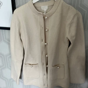 Beige kofta med pärlknappar från H&M - Supersöt beige kofta från H&M med rund halsringning och dekorativa pärlknappar framtill. Koftan har två små fickor framtill, båda med pärlknapp, och är stickad i mjukt material. Perfekt att slänga över en topp för en chill look.