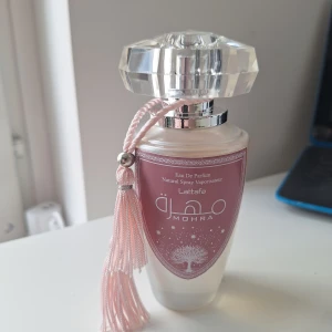 Mohra Silky Rose parfym från Lattafa 100 ml - Mohra Silky Rose från Lattafa är en elegant parfym i en frostad glasflaska med silverdetaljer och en stor, kristalliknande kork. Flaskan har en rosa etikett med trädmotiv och en dekorativ ljusrosa tofs. Doften har tydliga toner av ros, mysk, frukt, vanilj och tropiska inslag.(Tagit ca 2 ml. Locket glappar något)