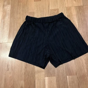 Svarta plisserade shorts med resår - Snygga svarta shorts med plisserat tyg och elastisk midja. De har en loose fit och är perfekta för en chill och trendig look. Materialet känns lätt och luftigt, vilket gör dem grymma för varmare dagar.