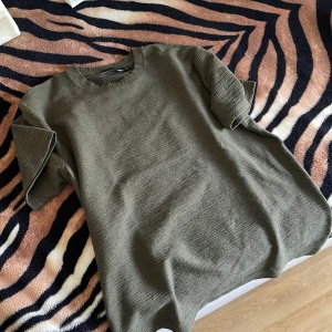 Olivgrön ribbad t-shirt - Snygg olivgrön t-shirt med ribbad struktur och rund halsringning. T-shirten har korta ärmar och är tillverkad i ett mjukt material som känns skönt mot huden. Perfekt för dig som gillar en enkel men stilren look. 10/10 skick 