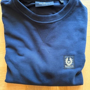 Mörkblå sweatshirt från Belstaff - Snygg mörkblå sweatshirt från Belstaff med rund hals och diskret logopatch på bröstet. Tröjan är i mjuk bomull och har långa ärmar samt ribbade muddar vid ärmslut och nederkant. Perfekt för en clean och stilren look.