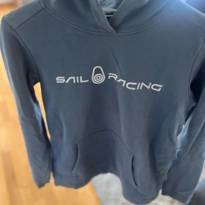 Blå hoodie från Sail Racing - Mörkblå hoodie från Sail Racing med vit logga framtill och på huvan. Klassisk känguruficka och långärmad modell. Perfekt för dig som gillar sportig och avslappnad stil. Mjuk och skön bomullskänsla.