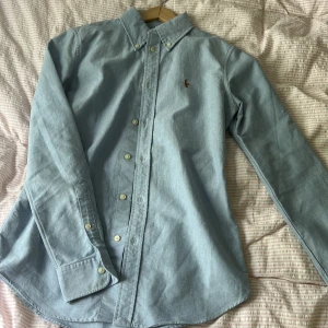 Ljusblå skjorta från Ralph Lauren XS - Snygg ljusblå skjorta från Ralph Lauren i bomull, custom fit. Klassisk krage, knäppning framtill och broderad polospelare på bröstet. Perfekt passform och stilren look, passar till allt. Långärmad modell med knappar vid ärmslut. Figursyd. 
