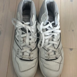 New Balance 650 vita sneakers - Snygga New Balance 650 sneakers i vitt med grå och svarta detaljer. Modellen är high-top med klassisk snörning och tydlig N-logga på sidan. Ovandelen är i skinn och textil, med perforerade partier för extra ventilation. Perfekta för dig som gillar retro basketstil.